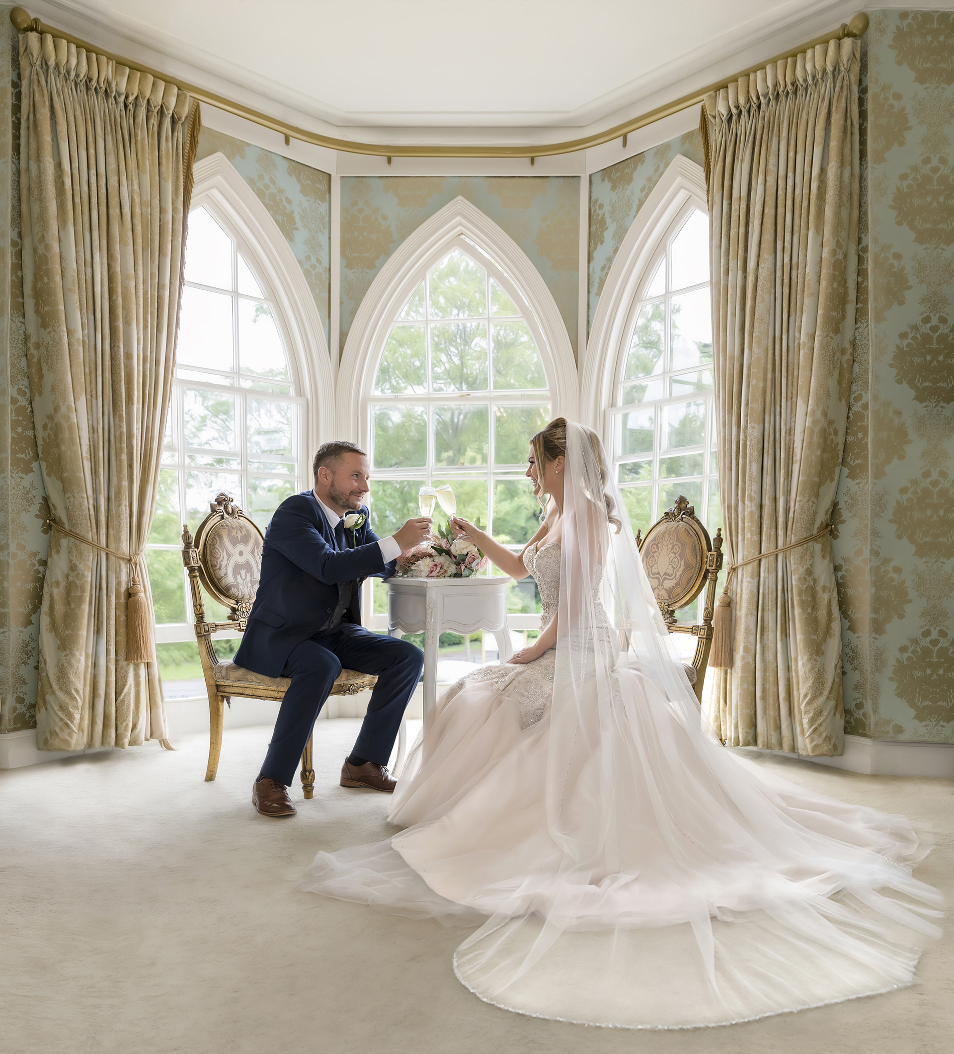 bride & groom siting in front ornate window, enjoying a glass of champaigne